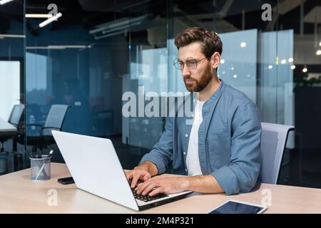 Serious young man freelancer, designer, IT specialist works concentratedly in the office, co-working space. Sitting at a table with a laptop. Stock Photo