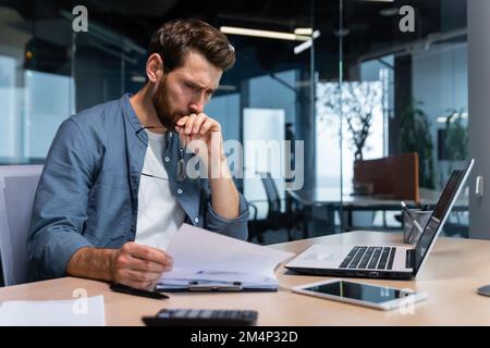 A serious young man accountant, financier, analyst, auditor sits in the office at the table. Thoughtfully holds glasses, looks, analyzes accounts, budget, works with documents and laptop. Stock Photo