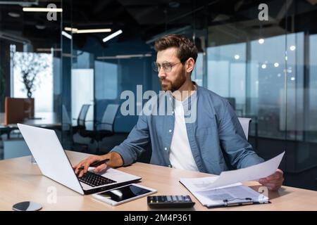 A serious young man accountant, financier, analyst, auditor sits in the office at the table. He holds documents and a pen in his hands, checks accounts, finances, types on a laptop. Stock Photo