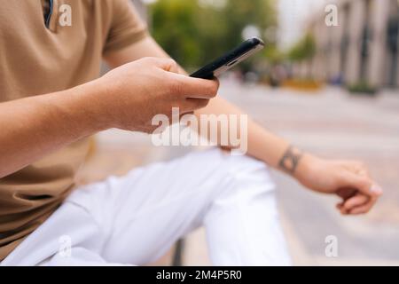 Close-up cropped shot of unrecognizable tattooed man sitting on bench using mobile phone looking to screen on city street. Stock Photo