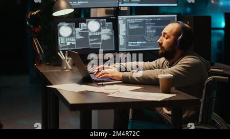 Young programmer typing source code in office, dealing with physical disability at night. Software engineer wheelchair user working late to develop new server database with html script. Stock Photo