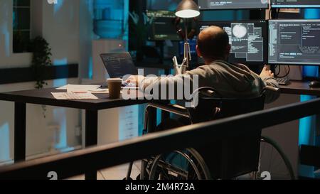 System engineer with impairment working on web programming, using laptop and multiple screens. Male IT coder with chronic disability in wheelchair developing new user interface. Stock Photo