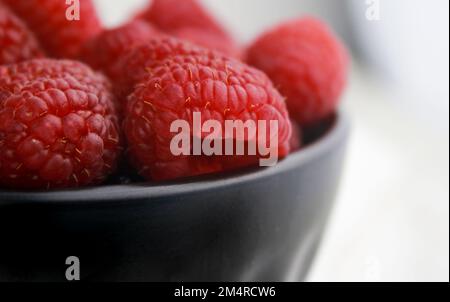 close up of fresh raspberries in dark bowl, blurred white background  Stock Photo