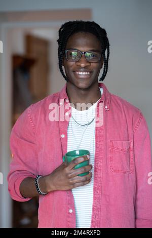 Happy young African man in glasses standing at home holding cup of tea or coffee and smiling Stock Photo