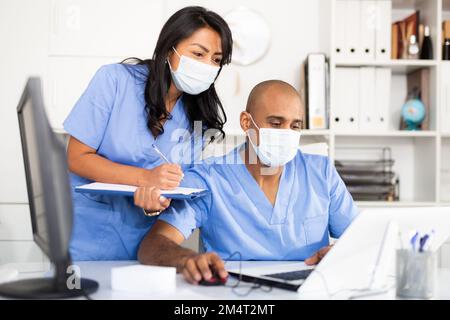 Two physicians in medical masks working with case histories on laptop Stock Photo