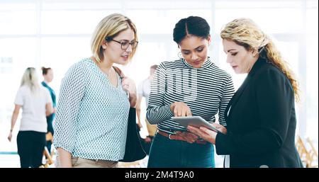Filling them in. three corporate businesswomen standing in a huddle with digital tablets in hand. Stock Photo