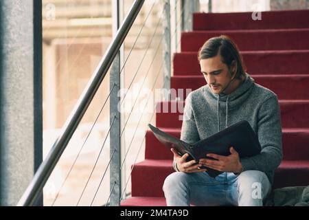 Preparing for exams. a handsome young university student studying while sitting on a staircase on campus. Stock Photo