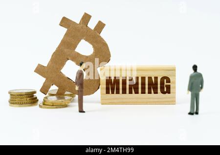 Cryptocurrency and business concept. On the white surface of the coin, the bitcoin symbol, miniature figures of businessmen and a wooden plate with th Stock Photo