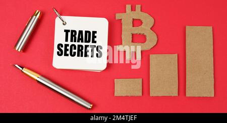 Business and bitcoin concept. On a red surface lie a bitcoin symbol, a graph, a pen and a notepad with the inscription - TRADE SECRETS Stock Photo