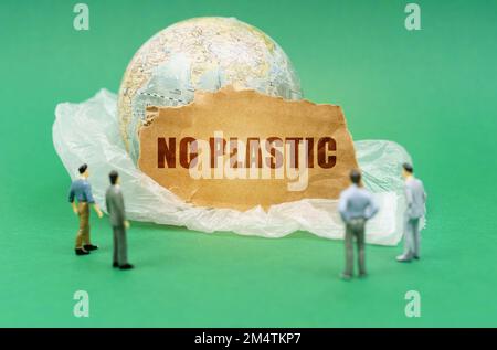 Ecological concept. On a green background on a plastic bag is a globe and a sign with the inscription - NO PLASTIC. Near out of focus figures of peopl Stock Photo