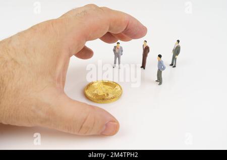 Cryptocurrency and business concept. On a white surface, bitcoin, miniature figures of people and a human hand that reaches for bitcoin. Stock Photo