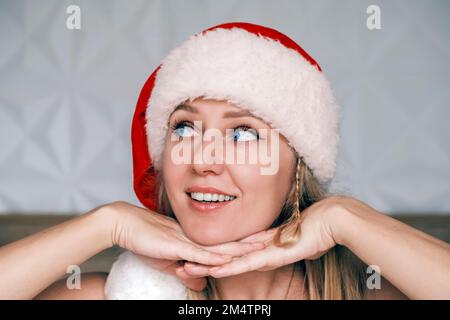 Smiling Santa girl wearing red hat is laying her chin on both hands looking up side. Portrait of beautiful young blue-eyed blonde woman in Santa's hat Stock Photo