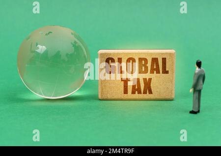 Business and finance concept. On the green surface is a globe and a figurine of a man who looks at a sign with the inscription - Global Tax Stock Photo