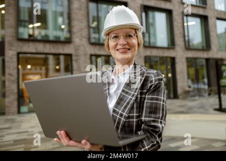 Senior female engineer in suit with white safety helmet working laptop on city building background Stock Photo