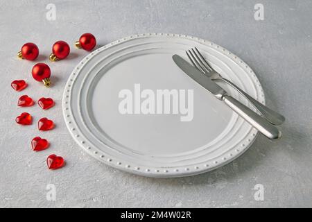 An empty white ceramic plate with cutlery surrounded by red christmas balls and hearts on a light concrete table Stock Photo