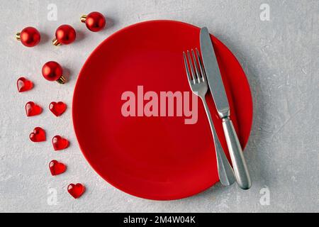 An empty red ceramic plate with cutlery surrounded by red christmas balls and hearts on a light concrete table Stock Photo