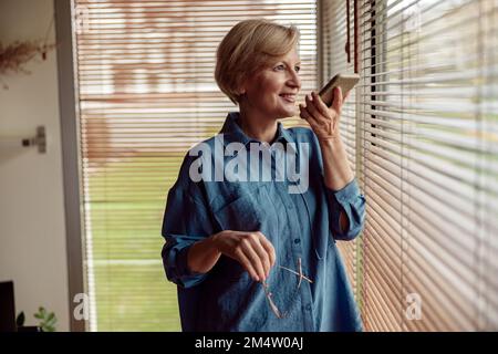 Middle aged woman recording audio message or using voicemail mobile function, standing near window Stock Photo