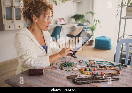 One woman young mature age at home making jewels and necklaces for online business activity. Female people sitting at the table in apartment doing bea Stock Photo