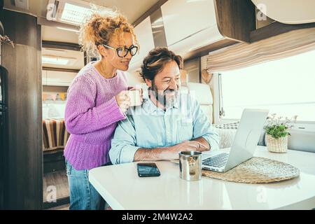 Happy couple man and woman working and using together a laptop computer inside a camper van planning holiday vacation destination and living vanlife l Stock Photo
