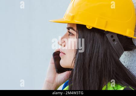 young long haired venezuelan caucasian engineer latina woman, wearing yellow safety helmet and vest, standing outdoors leaning against a wall, on a ph Stock Photo