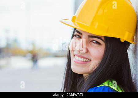 closeup young caucasian latin woman of venezuelan ethnicity engineer happy outdoors leaning on wall smiling and looking at camera, wears yellow hardha Stock Photo