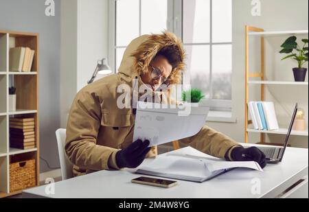 Man in a winter clothes looking at heating bills while sitting at his desk in his cold house Stock Photo