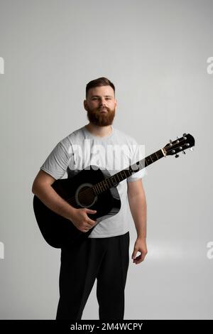 Male musician playing on acoustic guitar. Portraite of bearded handsome musician holding a guitar in a white room. Guitarist, artist, composer, rocker Stock Photo