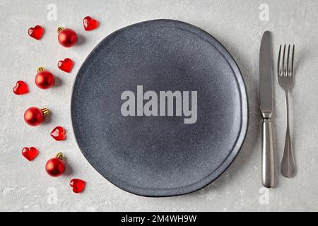 An empty gray ceramic plate with cutlery surrounded by red christmas balls and hearts on a light concrete table Stock Photo