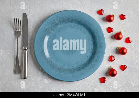 An empty blue ceramic plate with cutlery surrounded by red christmas balls and hearts on a light concrete table Stock Photo