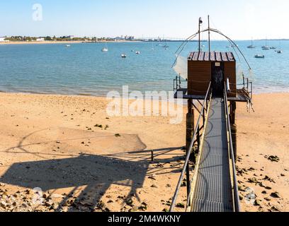 A wooden fishing cabin with a large square lift net called 'carrelet' casts its shadow on the beach at low tide, facing the Saint-Nazaire bridge. Stock Photo