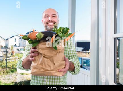 Smiling man holding paper bag full of vegetables at doorway Stock Photo