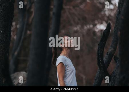 Thoughtful woman standing amidst burnt trees in forest Stock Photo