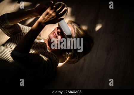 Mature woman wearing VR glasses and holding light bulb lying at home Stock Photo