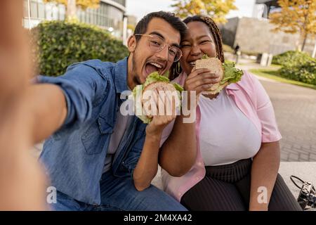 Happy young couple eating sandwich and taking selfie at footpath Stock Photo