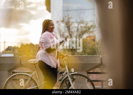 Happy young woman standing with bicycle and using smart phone Stock Photo