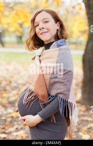 Smiling pregnant woman with shawl standing in autumn park Stock Photo