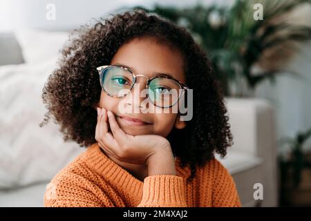Smiling girl wearing eyeglasses sitting with hand on chin at home Stock Photo