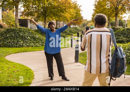 Man photographing woman standing with arms raised at park Stock Photo