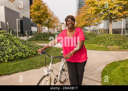 Smiling senior woman wearing headphones wheeling bicycle at park Stock Photo