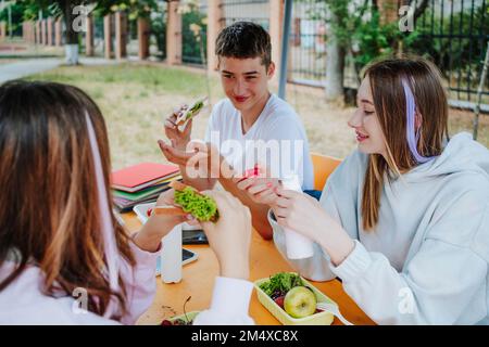 Girls with boy having lunch sitting at table in schoolyard Stock Photo