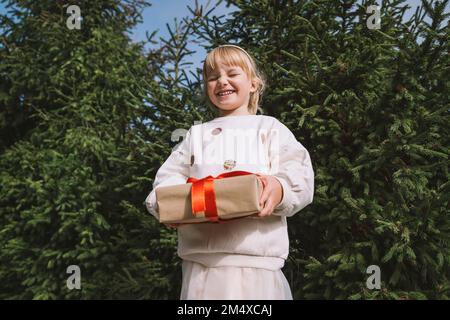 Smiling girl with gift box standing in front of fir tree Stock Photo