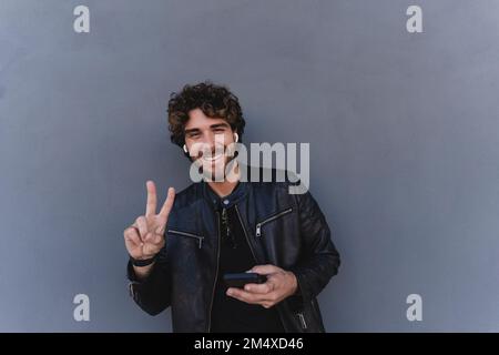Happy man holding mobile phone gesturing peace sign leaning on gray wall Stock Photo
