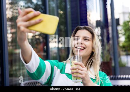 Happy young woman holding drinking glass taking selfie through mobile phone Stock Photo