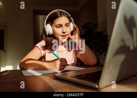 Girl wearing wireless headphones studying on laptop at desk Stock Photo
