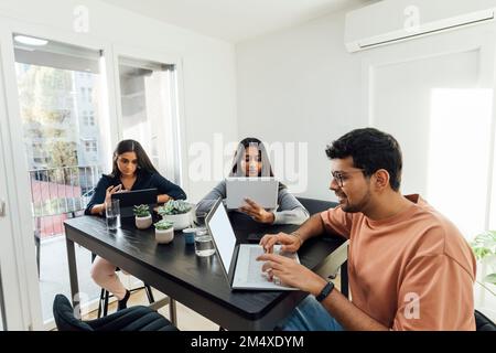 Smiling man using laptop in front of women studying through tablet PCs on table in living room Stock Photo