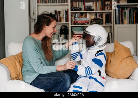 Happy mother looking at daughter wearing space costume on sofa Stock Photo