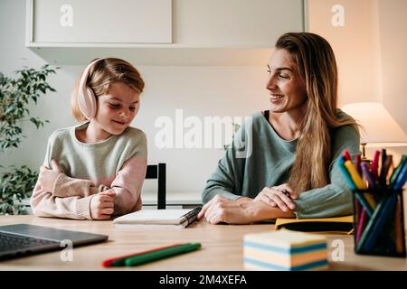 Happy mother looking at daughter sitting with notebook at table Stock Photo