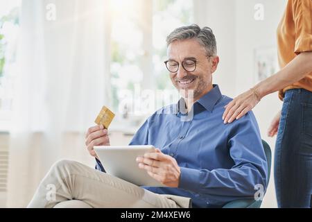 Happy man holding credit card doing online shopping over tablet PC with hand of woman on shoulder at home Stock Photo