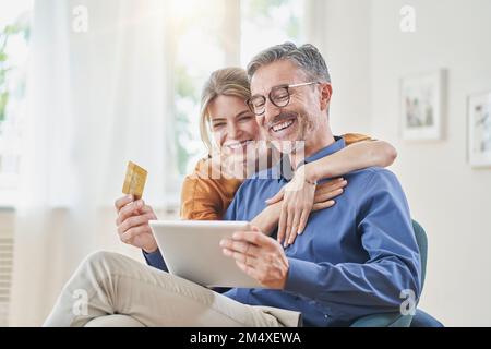 Smiling woman embracing man with credit card doing online shopping through tablet PC at home Stock Photo