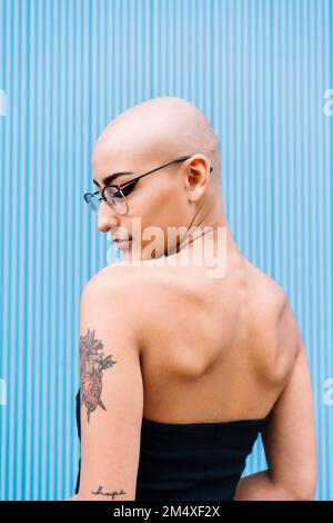 Woman with shaved head wearing eyeglasses in front of blue wall Stock Photo